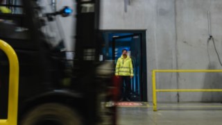 Um homem com vestuário de segurança sai de uma porta para o pavilhão de uma fábrica. Passa um empilhador. Um homem com vestuário de segurança sai de uma porta para o pavilhão de uma fábrica. Passa um empilhador.