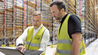 Os processos de carregamento em paralelo podem dar origem a picos de carga indesejados. O connect:charger da Linde pode ajudar. Dois homens com coletes de segurança amarelos estão num armazém de estanterias altas a olhar para um computador portátil.