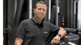 Tibor Sevenheck da Future Warehouse Team da Arvato. Tibor Sevenheck encosta-se a um empilhador para corredores estreitos e sorri para a câmara.