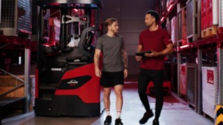 Duas pessoas com roupa desportiva estão em frente a um empilhador retrátil Ri16 da Linde MH; uma delas tem um tablet na mão.