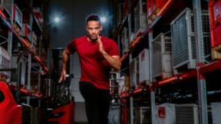 Um homem, vestido com roupa desportiva, corre entre as estanterias do armazém em direção à câmara.