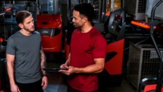 Dois homens, vestidos com roupa desportiva, estão num armazém a conversar.